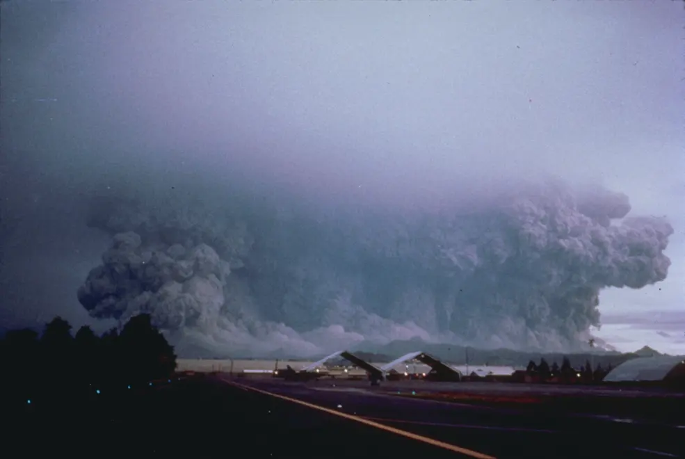 A huge gray cloud of ash billowing up and spreading out behind an airfield, under a dark sky