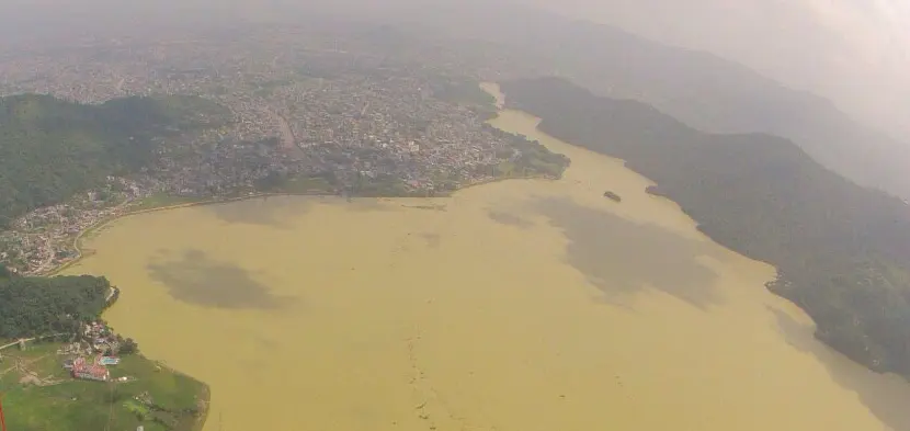 An aerial view of a large lake with muddy brown water, surrounded by green forested hills on one side and a town on the other