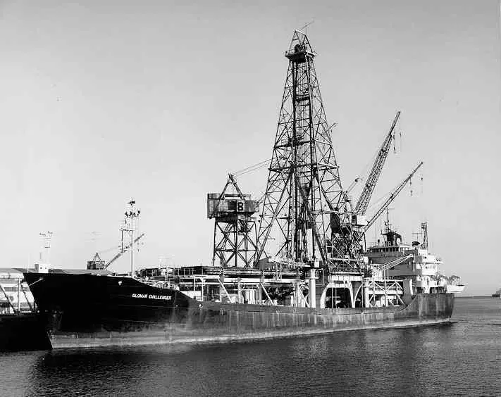A black-and-white photograph of a ship in harbor with a tall steel drilling tower in the middle of its deck