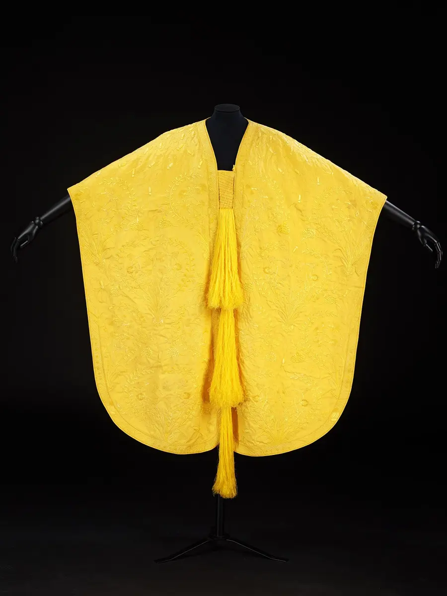 A bright golden-yellow embroidered cape with long tassels, displayed on a stand against a black background