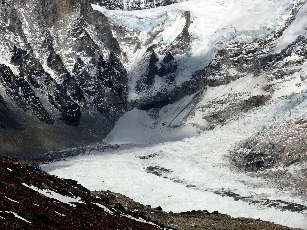 A wide, jumbled river of white ice blocks and crevasses pouring down between dark rocky mountainsides