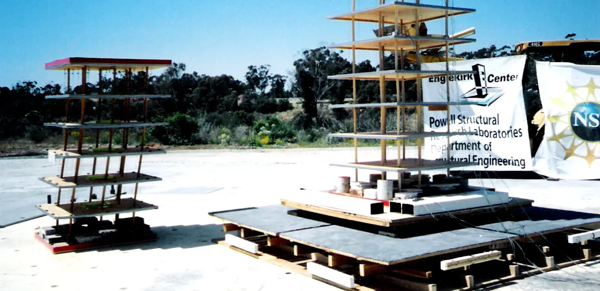 Two small model buildings of thin posts and flat floors, several storeys tall, standing side by side on the platform of an outdoor shake table