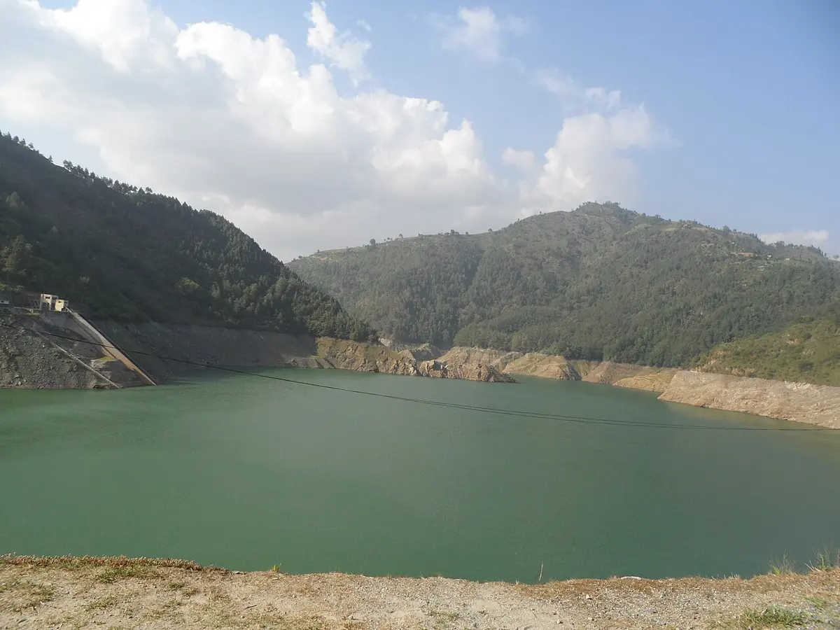 A green reservoir lake between forested hills, with a band of bare rock and soil along the shore above the water