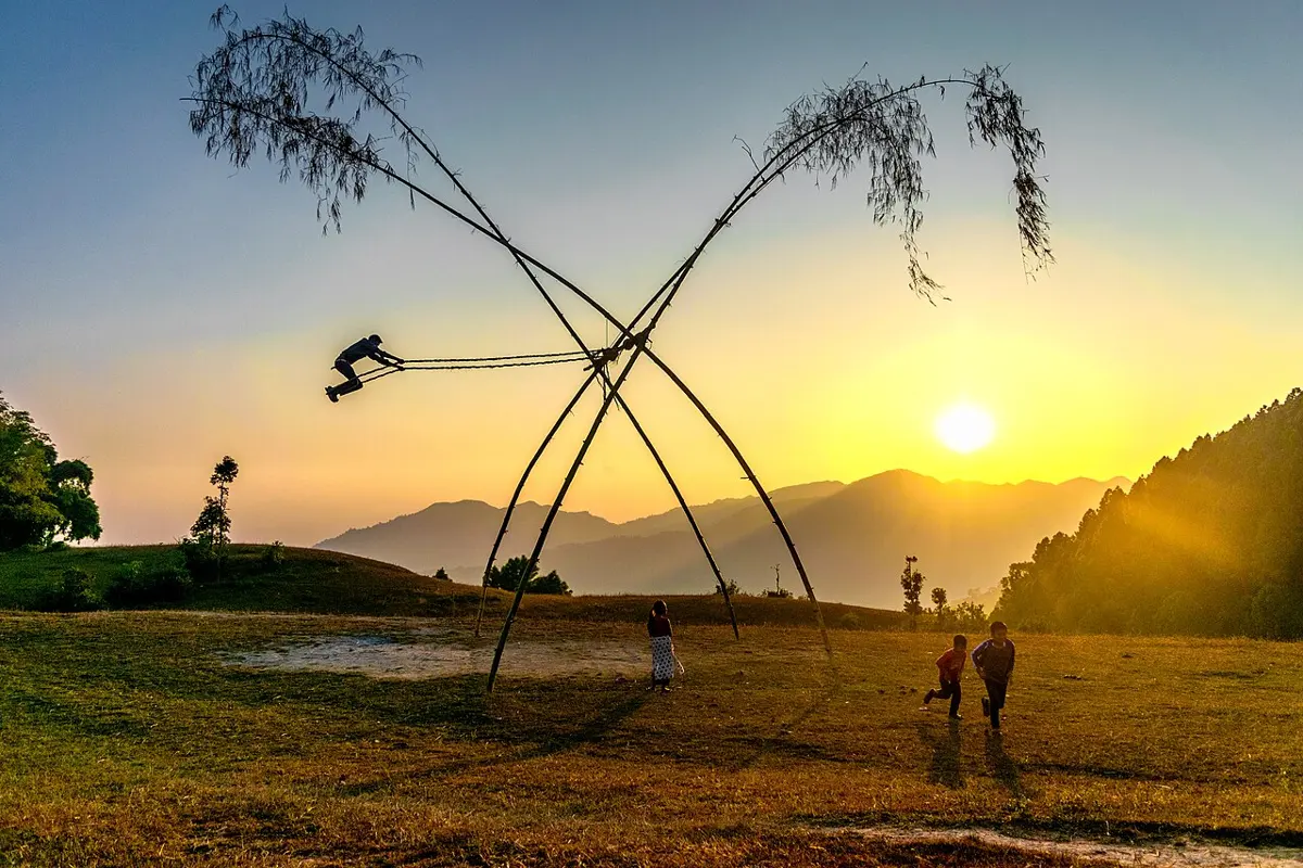 Four tall bamboo poles, their leafy tops left on, leaning together in two crossed pairs; a child standing on the swing's seat flies high out to one side, against a sunset over hills