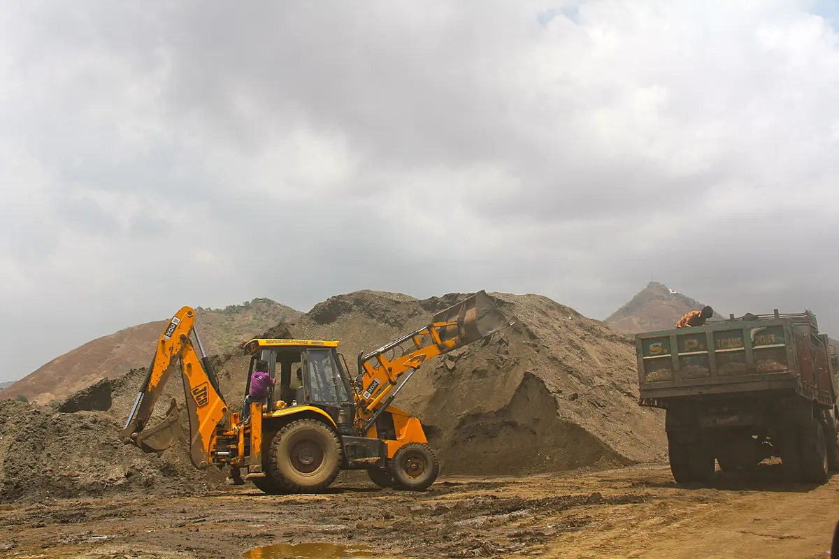 A yellow backhoe loader tipping a bucket of sand into a tipper truck beside tall heaps of sand under a cloudy sky