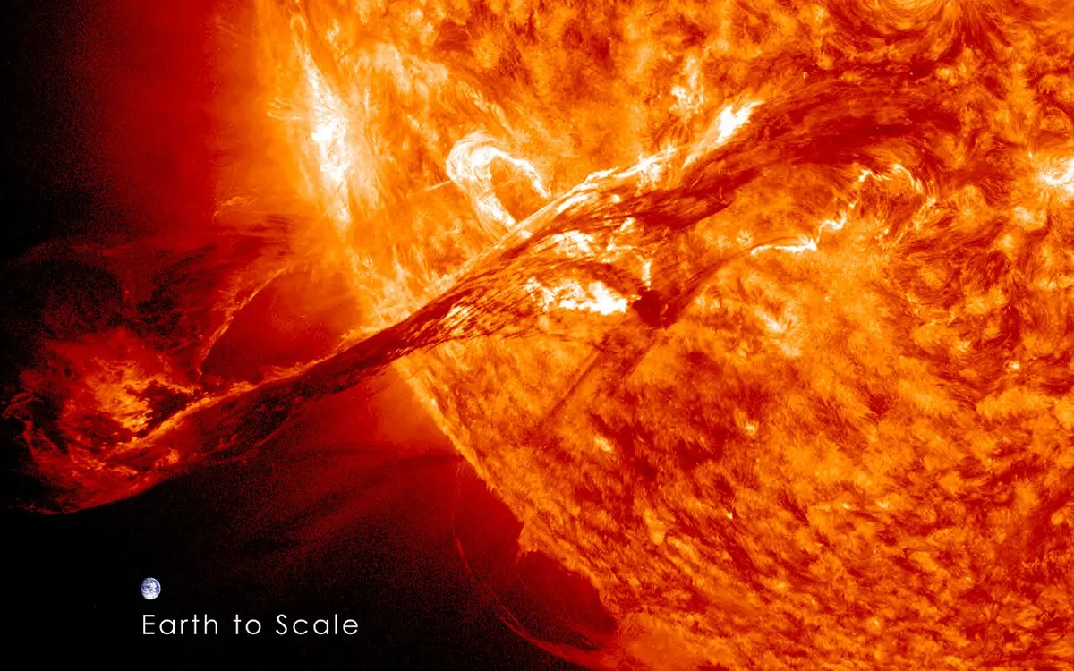 A huge loop of glowing orange gas bursting out from the edge of the Sun into space, with a tiny picture of the Earth added beside it for size