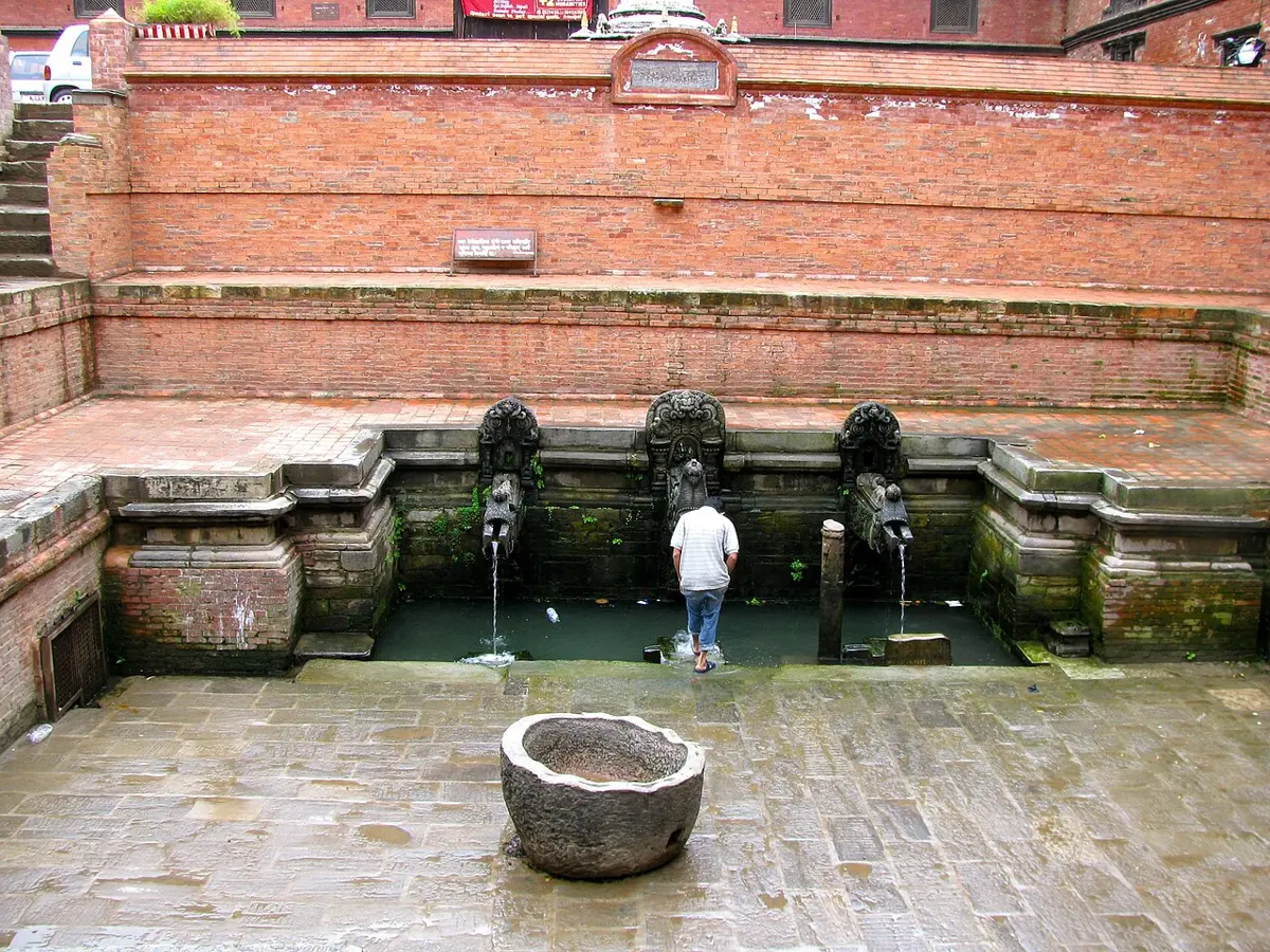 A sunken brick courtyard with three carved stone water spouts in a wall at the bottom, a man standing by the pool below them, and a round stone basin in the paved floor in front