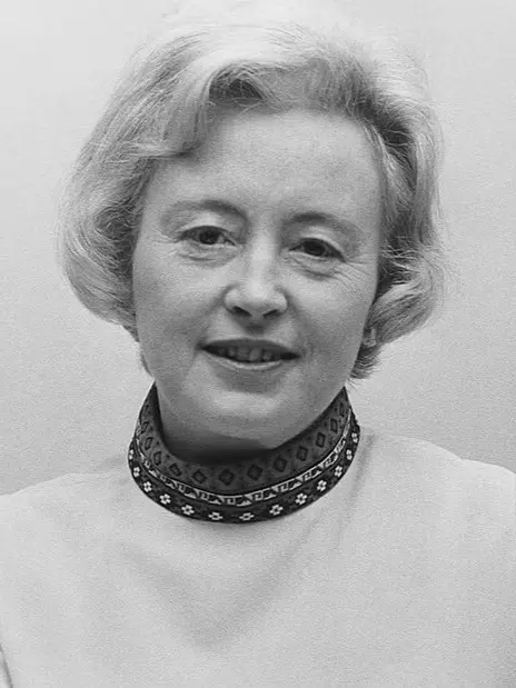 A black-and-white photograph of a smiling woman with short, wavy hair, wearing a pale jumper with a patterned collar