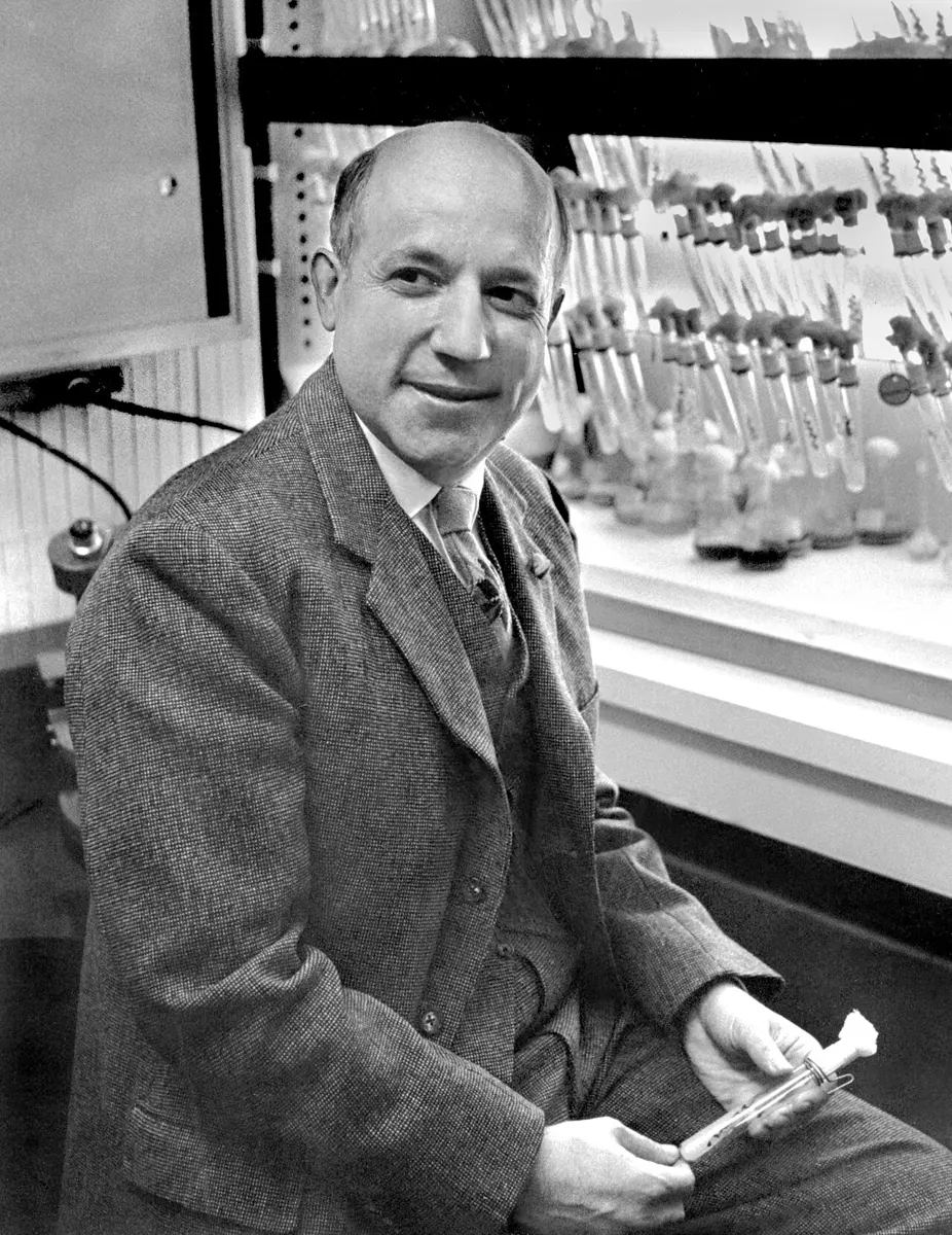 A black-and-white photograph of a smiling man in a tweed jacket, seated in a laboratory in front of racks of glass flasks and tubes, holding a test tube