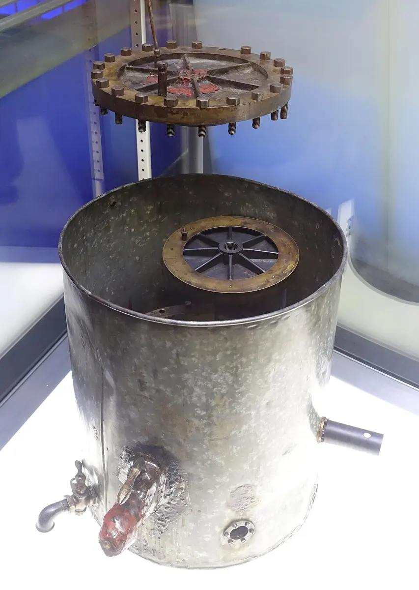 An open metal drum with a tap, a side tube and a small round window, its heavy bolted lid hanging above it, in a museum case