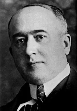 A black-and-white portrait of a balding man in a suit, white collar and striped tie