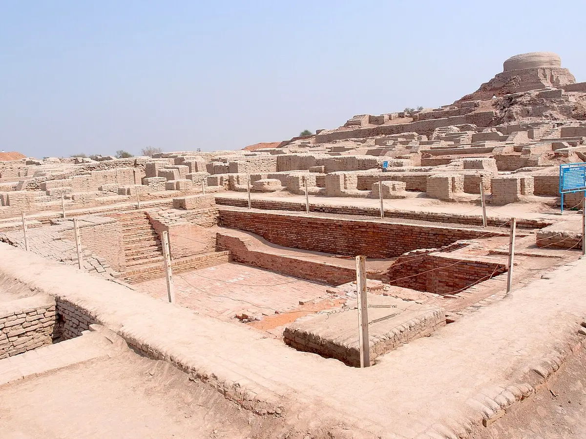 The ruins of a brick city under a clear sky: low walls of pale baked bricks, a large rectangular sunken pool with steps down into it, and a mound with a round brick dome in the distance