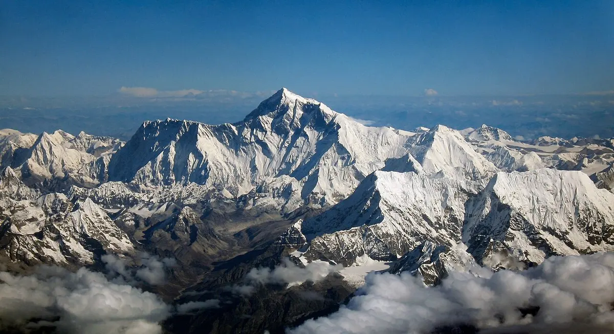 Mount Everest from the air: a snowy pyramid standing above a sea of white peaks and clouds, under a blue sky