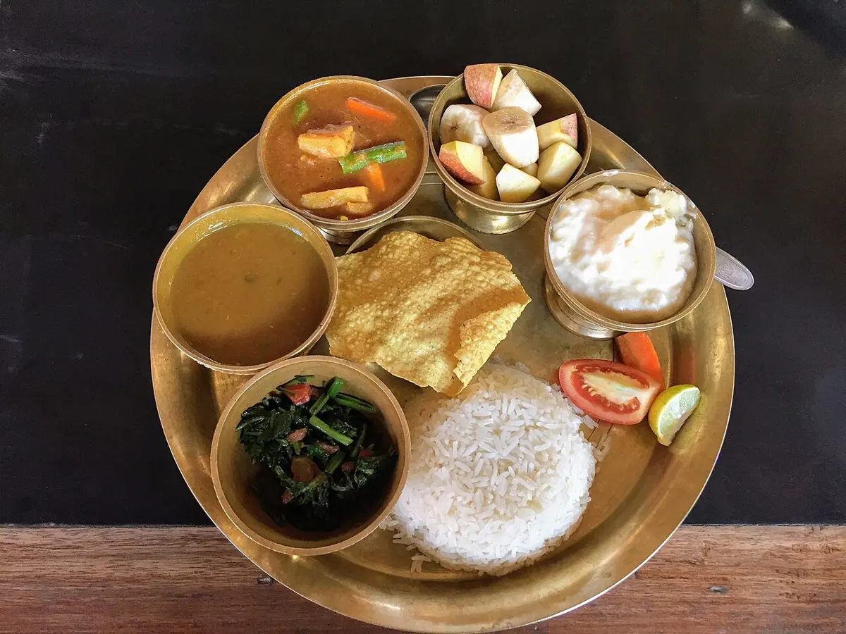 A brass plate with a mound of white rice, a papad, and small bowls of dal, green saag, vegetable curry, yoghurt and cut fruit, with slices of tomato and lime