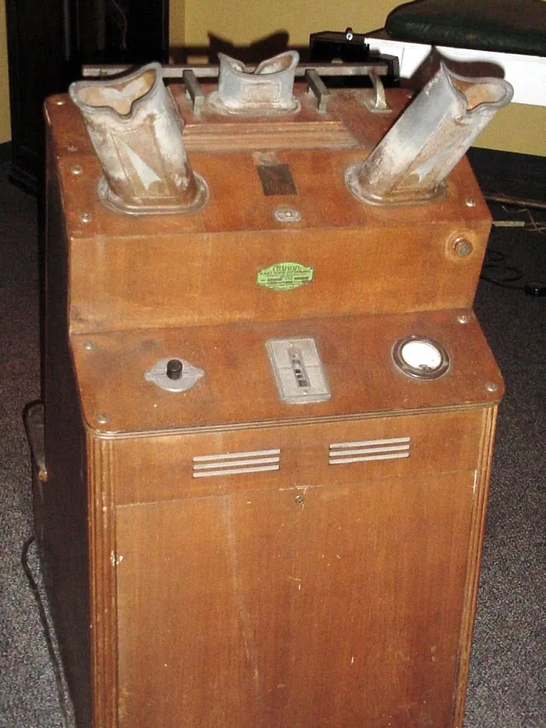 A waist-high wooden cabinet with three metal viewing eyepieces on top, a switch, a timer and a dial