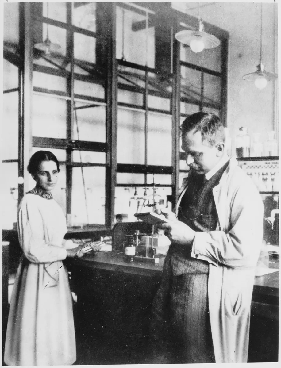 An old black-and-white photograph of a young woman in a long light dress and a man in a lab coat reading a notebook, in a laboratory with tall windows