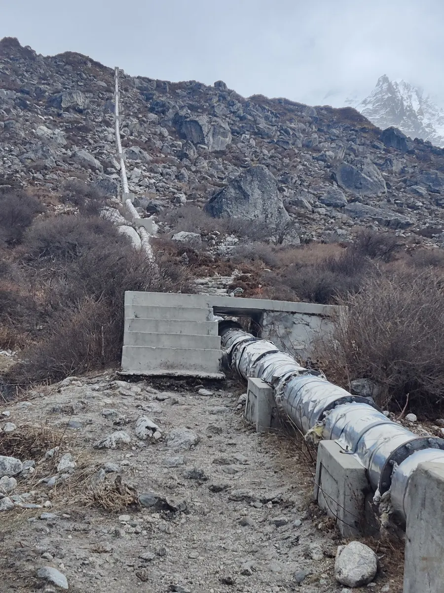A metal pipe on concrete supports leaving a concrete tank and running down a stony slope, with rocky mountains behind