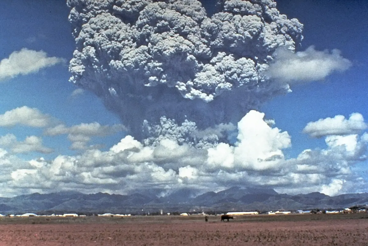 A huge gray column of volcanic ash and gas rising high above a range of mountains and white clouds, seen across a flat, bare field