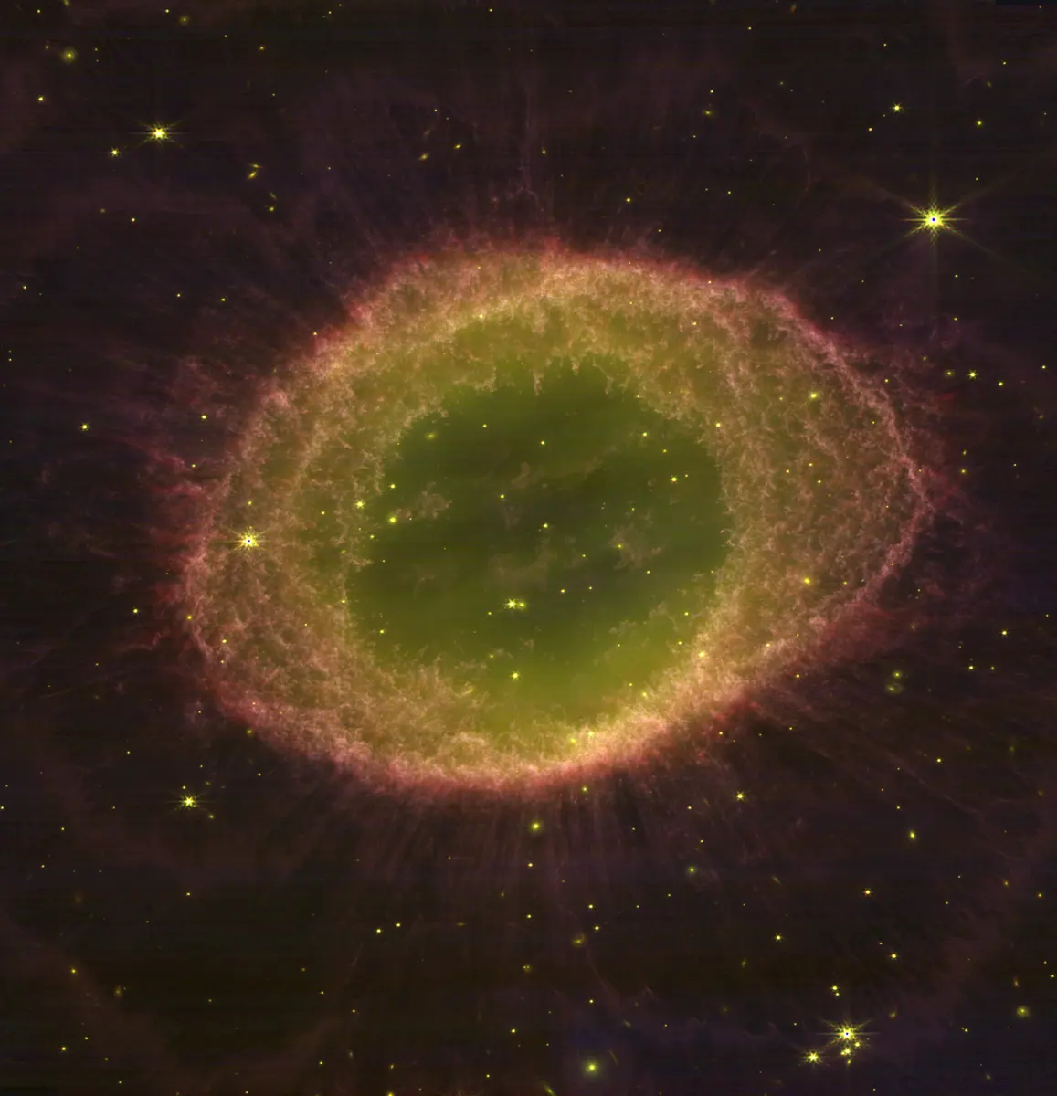 A glowing oval ring of gas, pinkish-orange on the outside and greenish inside, with faint streaks spreading outwards, against a dark sky scattered with stars