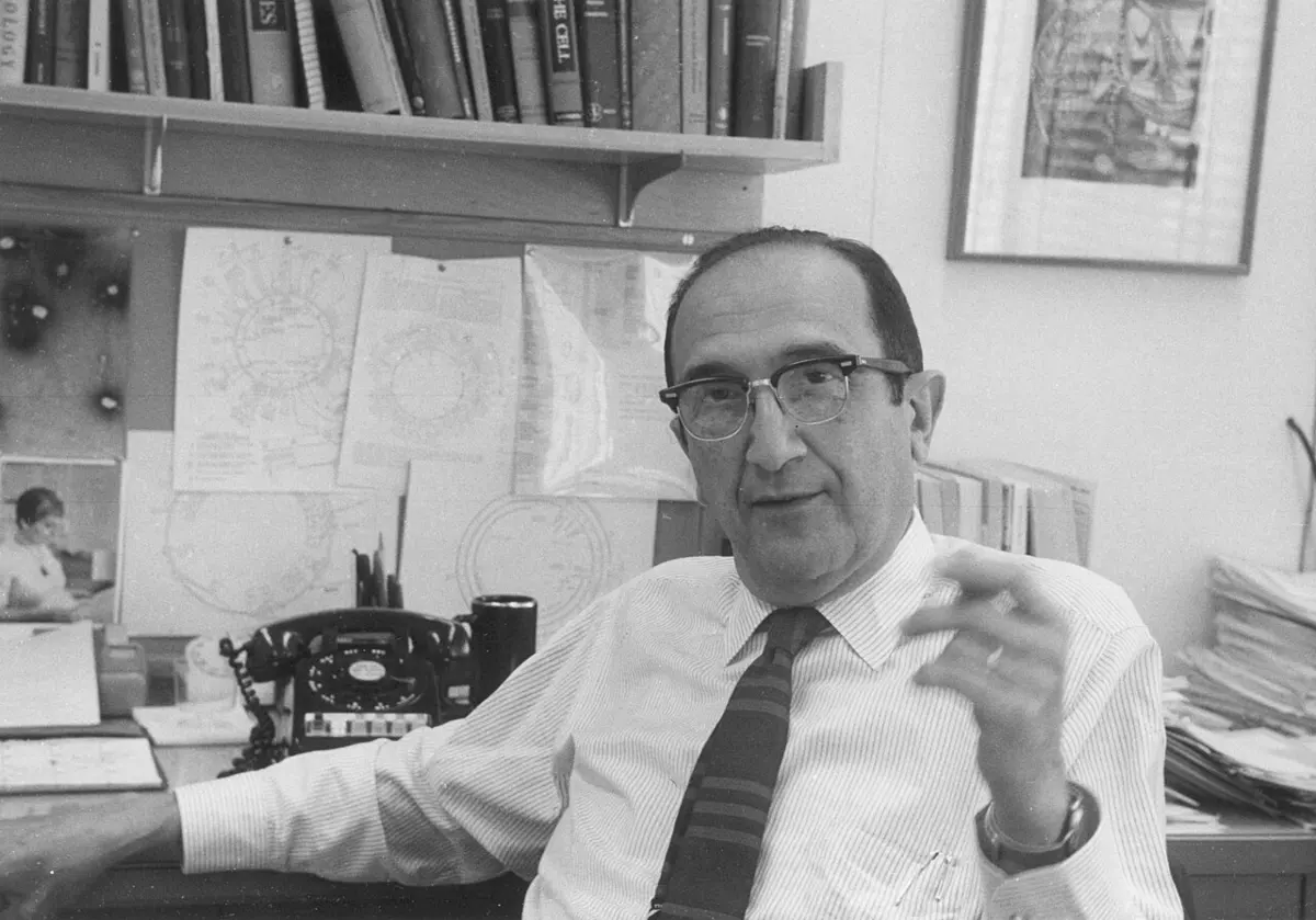 A black-and-white photograph of a man in glasses, a white shirt and a striped tie, talking at his desk in an office with bookshelves, a telephone and diagrams pinned to the wall