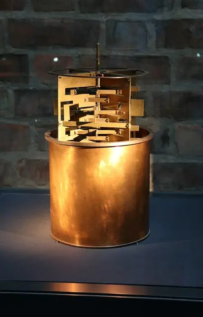 A copper can, open at the top, with a brass paddle wheel and its fixed vanes standing half out of it, on a museum display stand