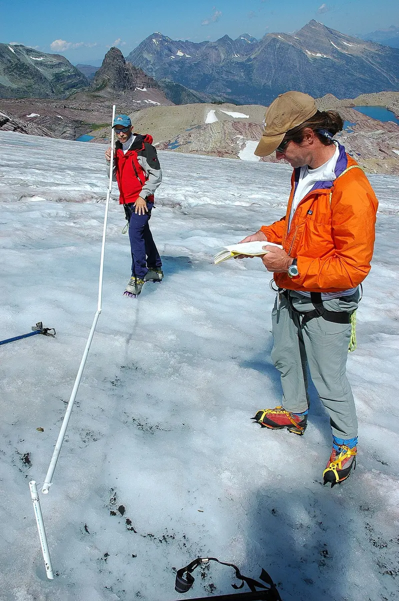 Two people in climbing gear standing on the white, gritty surface of a glacier. One holds up a long white pole made of jointed sections; the other reads a notebook. A short white tube sticks out of the ice at their feet