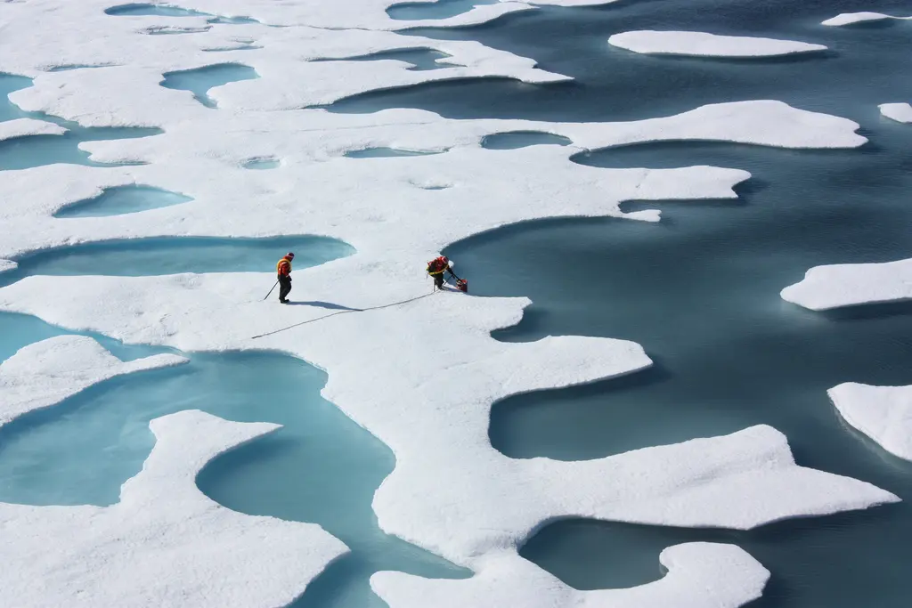 Two people in red jackets walking on white sea ice dotted with ponds of blue-green meltwater, with dark open sea on the right