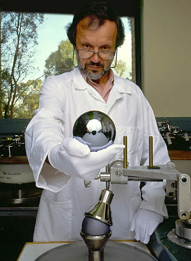 A man in a white laboratory coat and white gloves holding out a shiny, mirror-like ball the size of a large orange
