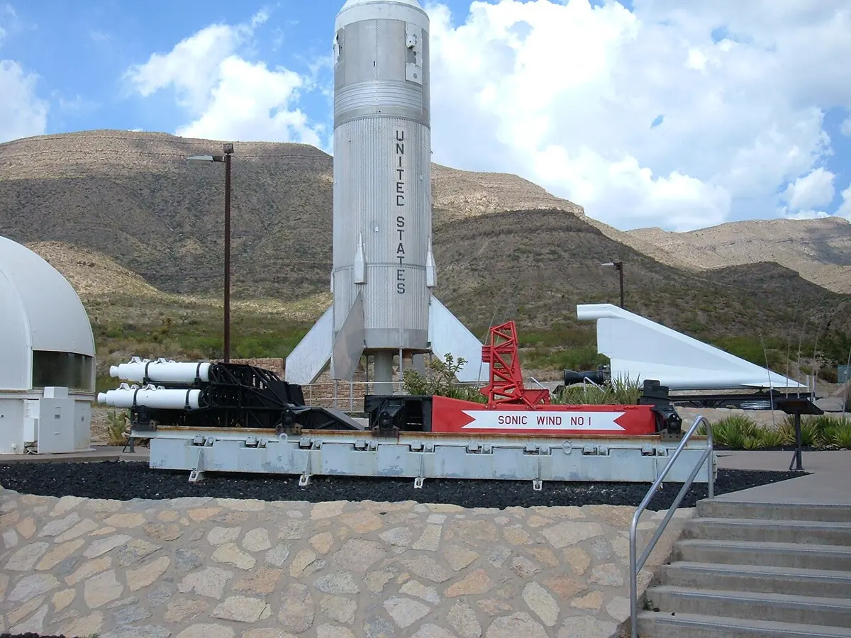 A rocket sled on display outdoors: a black frame with white rocket motors at the back and a red seat section with the sign Sonic Wind No 1, in front of a tall white rocket and desert hills