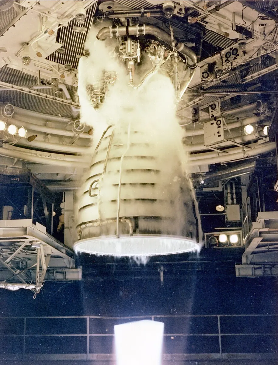 A large bell-shaped rocket engine hanging in a test stand, firing downwards; white vapor around the nozzle, and below it an almost invisible exhaust with a glowing patch