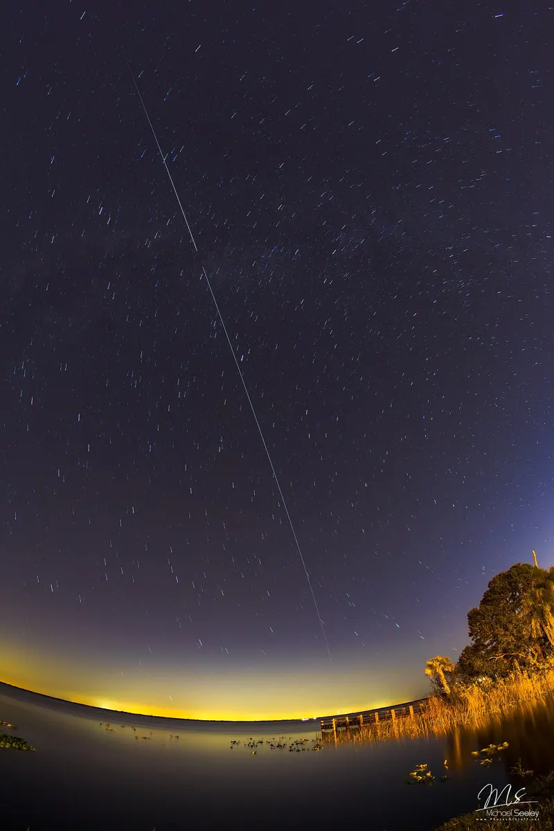A starry night sky over a lake, with a long, straight, thin streak of light running from high in the sky down towards the glowing horizon