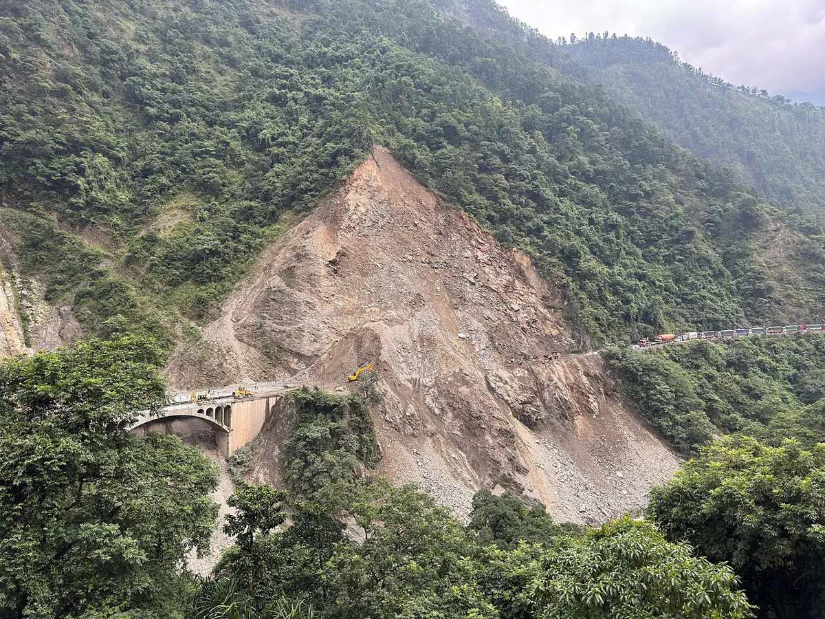 A steep hillside of pale, bare rock and rubble, a scar in the middle of green forest, running down to a road and a bridge in the valley