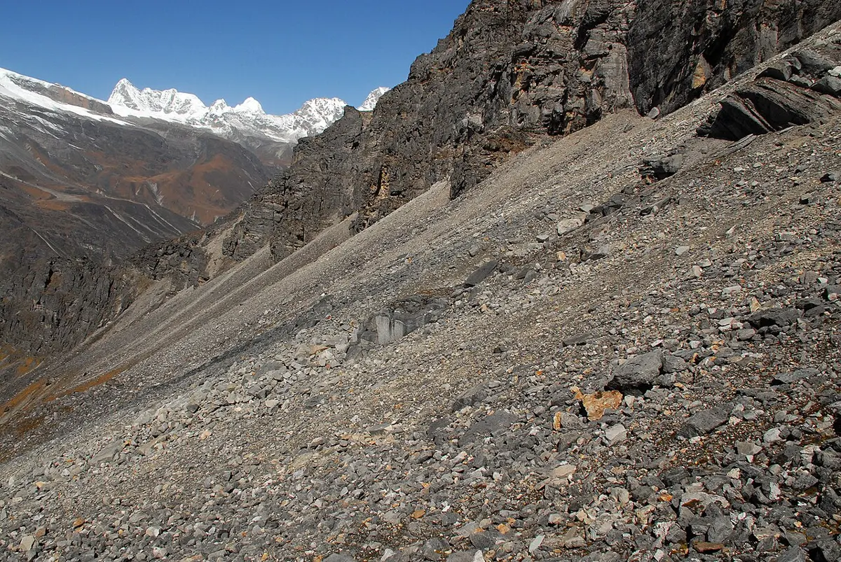 A steep slope of loose gray stones of all sizes below dark, cracked cliffs, with snowy peaks in the distance under a blue sky