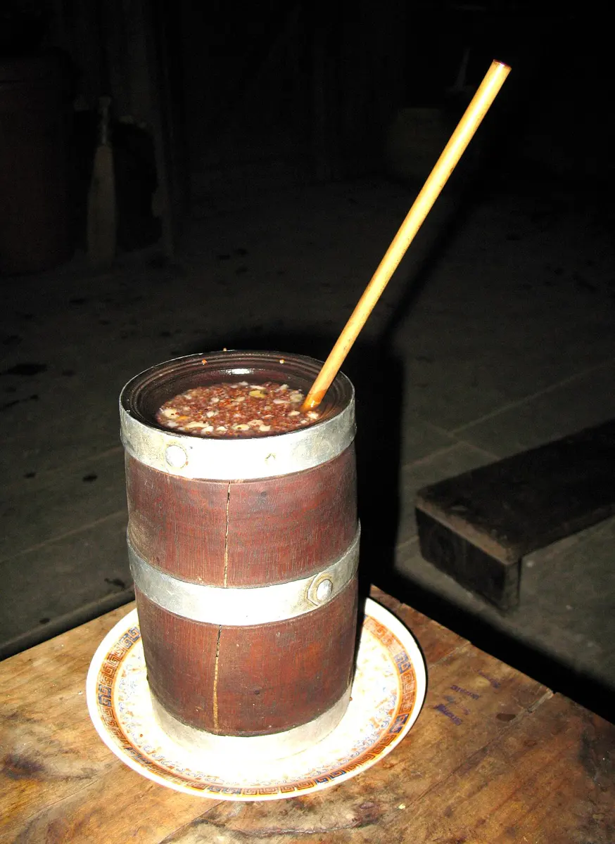 A small wooden mug bound with metal bands, full of fermented millet grains, with a bamboo straw, on a saucer