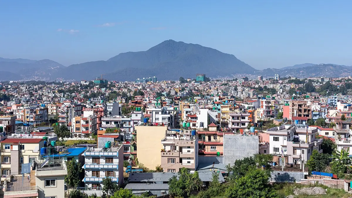 A dense city of many-storeyed concrete houses, white, pink and yellow, filling a flat valley floor right up to the foot of a large forested hill under a blue sky