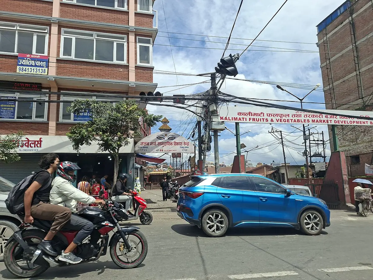 भिडभाड भएको सडकमा कालीमाटी फलफूल तथा तरकारी बजारको गेट, छेउबाट मोटरसाइकल र कार गुडिरहेका