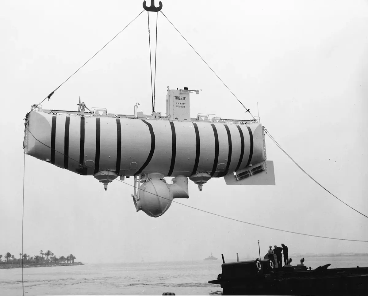 A long cylindrical craft with a small ball-shaped cabin underneath, hanging from a crane over the water