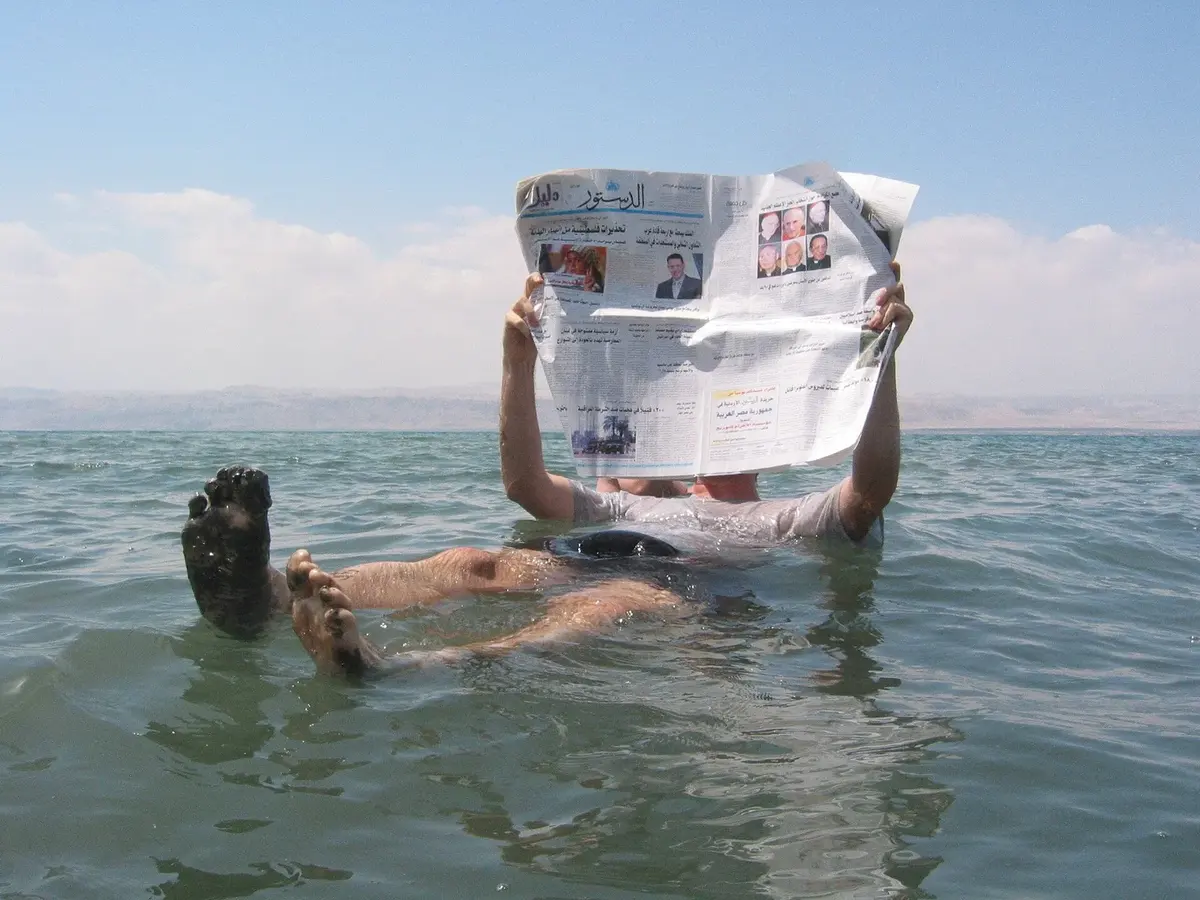 A man floating on his back in the Dead Sea, holding up a newspaper to read, with his feet sticking out of the water