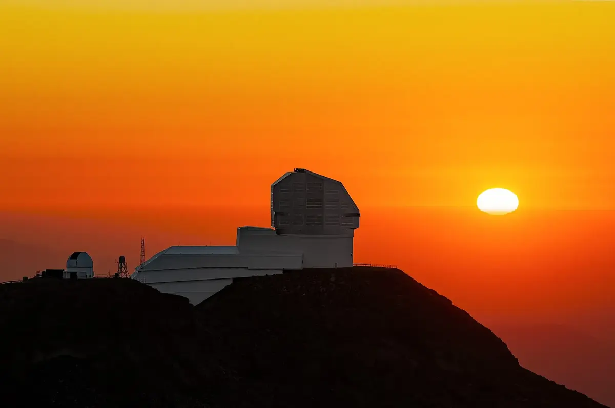 The setting sun, squashed into an oval with ragged layers, just above the horizon behind an observatory