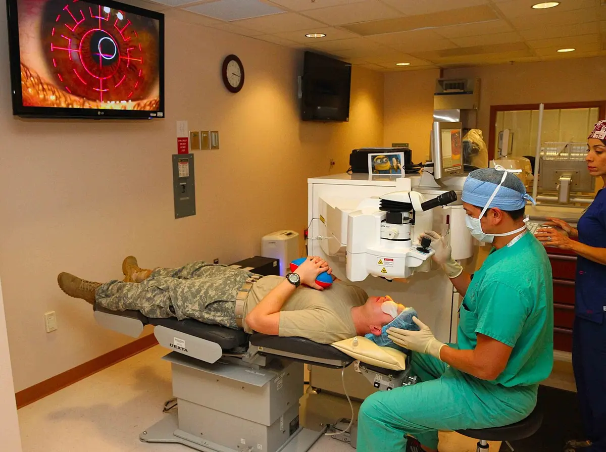 A patient lies on a reclining bed with his head under a large white laser machine; a surgeon in green scrubs looks into its microscope, and a screen on the wall shows the patient's eye, magnified