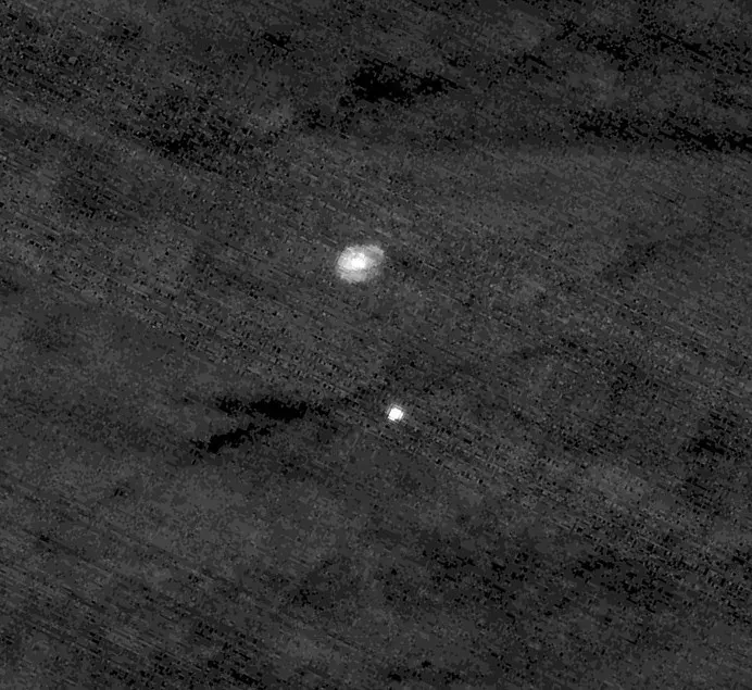 Seen from far above, a small white parachute and, below it, a bright dot: the capsule carrying the rover, against the blurred gray ground of Mars