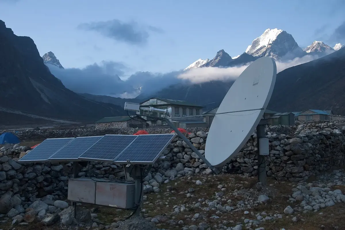 A satellite dish and solar panels inside a stone wall in a high Himalayan village, with snowy mountains behind