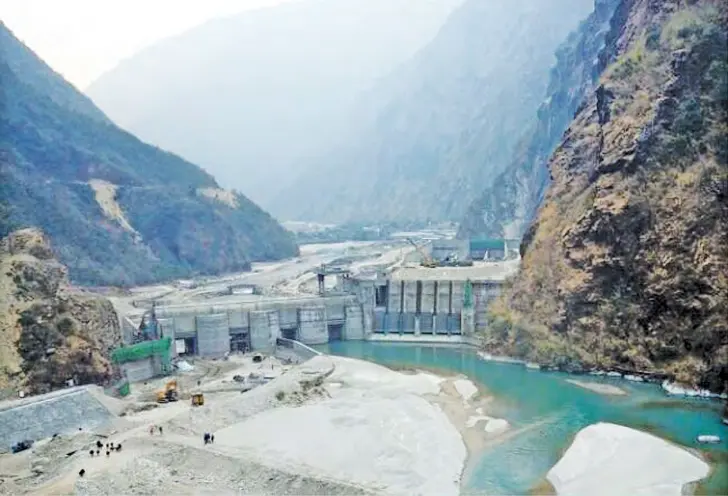 A concrete dam with tall gates across a narrow valley between steep rocky mountains, with green-blue water pooled in front of it and workers and machines on the riverbed