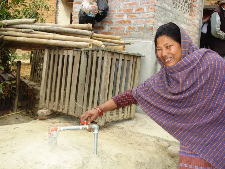 A smiling Nepali woman in a shawl turns the tap on a gas pipe that comes out of the ground in her yard