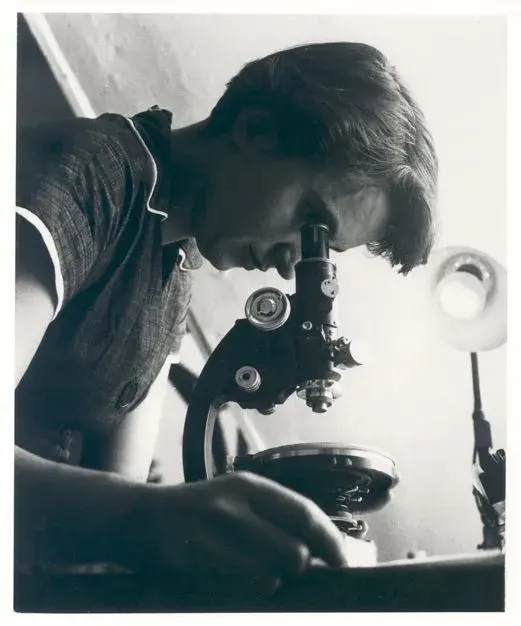 A black and white photo of a young woman looking into a microscope