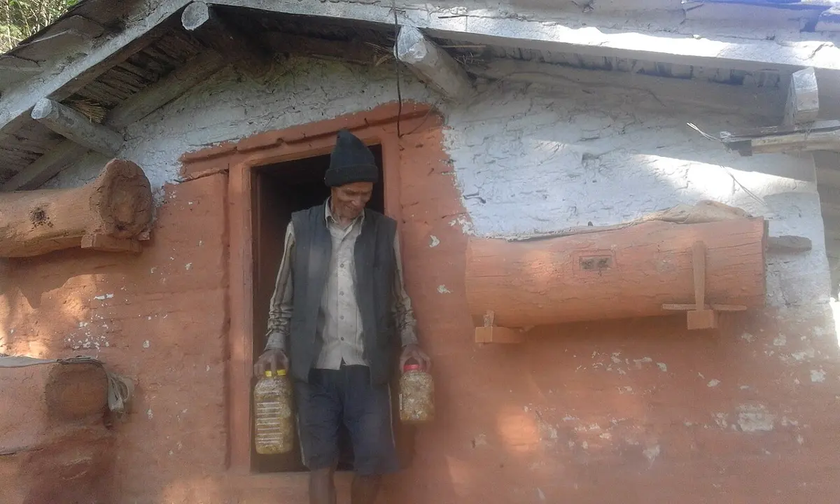 A smiling old man in a woolen cap stands in the doorway of a red mud house holding two plastic jars of honey; hollow logs with small entrance holes are fixed to the wall on both sides