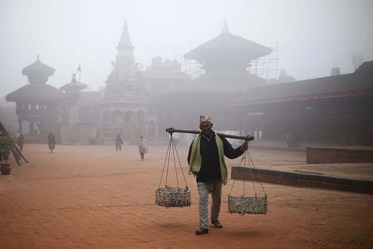 Temples of Bhaktapur Durbar Square half hidden in gray fog, with a man carrying two baskets on a pole across the brick square