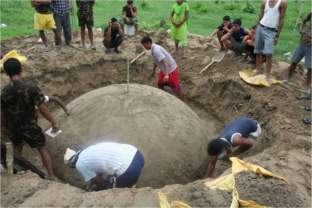 People building a biogas plant in a deep round pit; a big dome of smooth soil fills the pit, with a stick standing on top