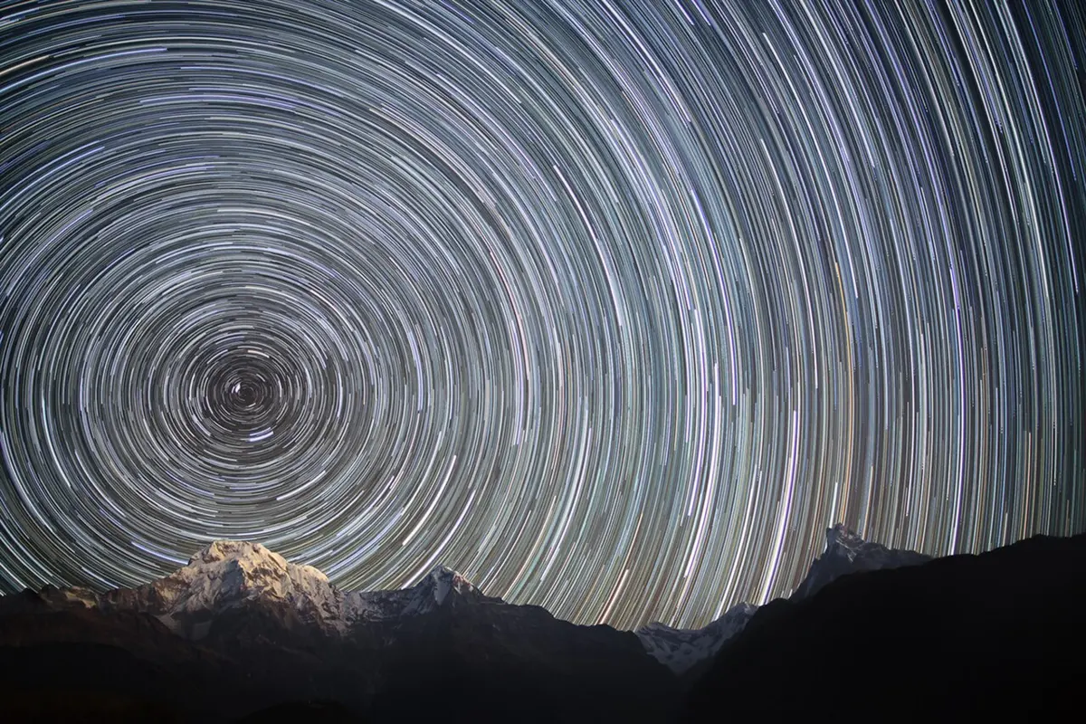 Circular streaks of starlight curving around one point in a dark night sky above snowy mountains