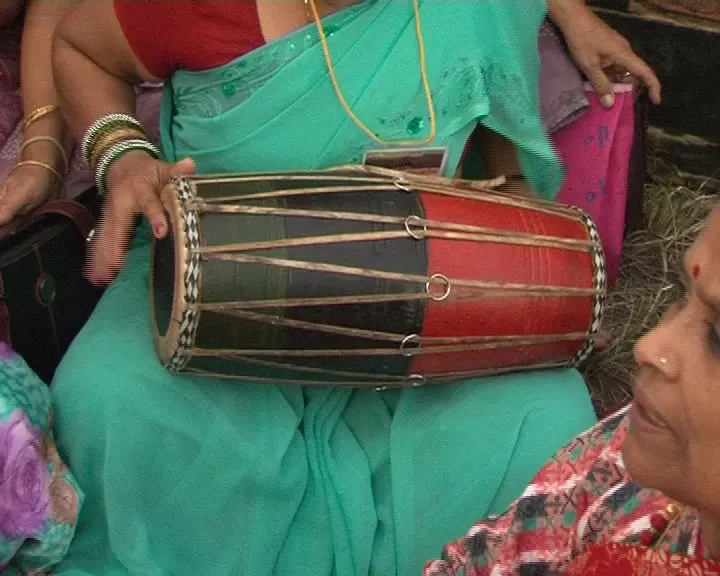 A woman in a green sari holding a madal, a barrel-shaped drum painted red and black, with drum skins at both ends tied with straps
